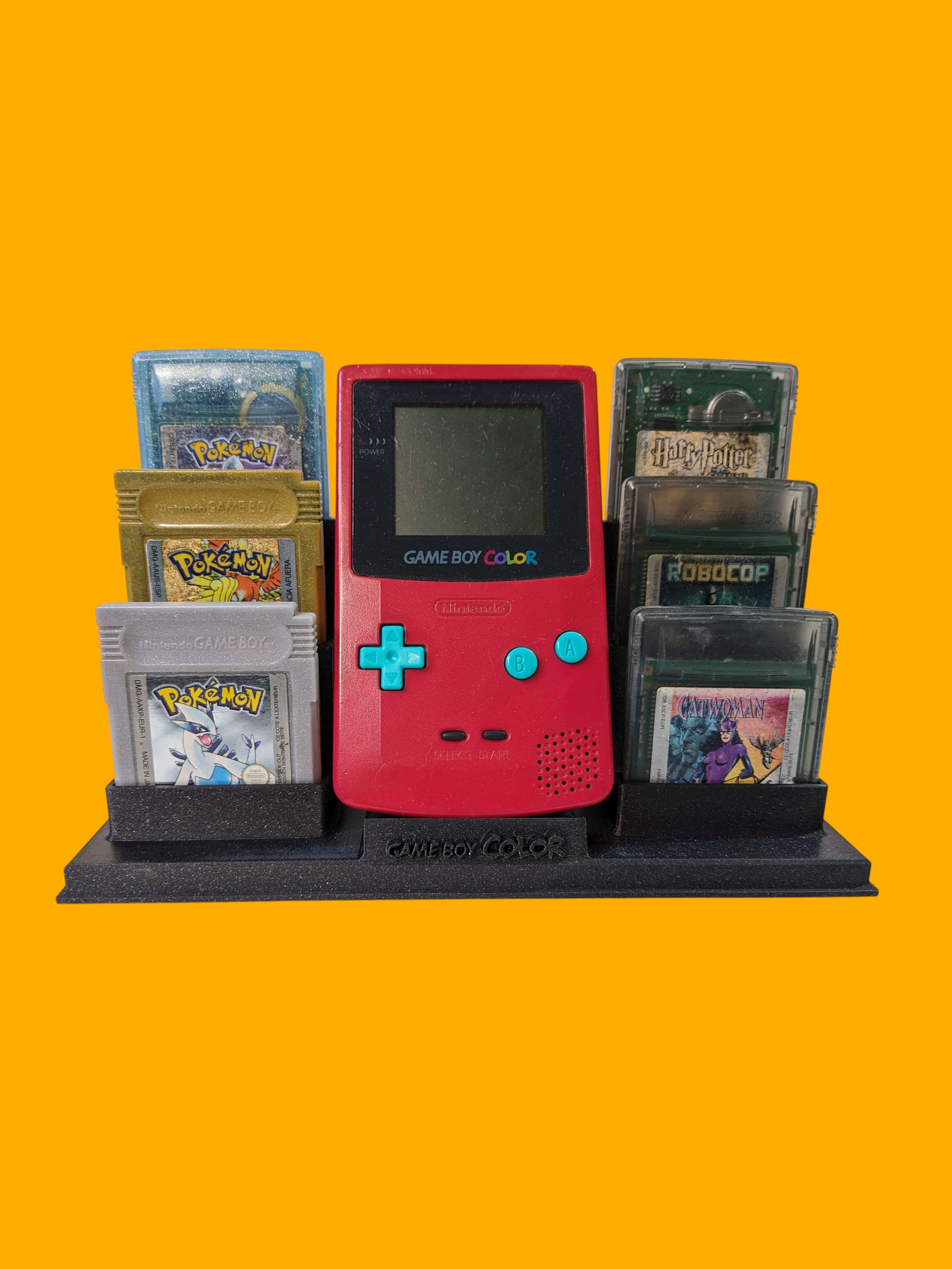 A bright red Game Boy Color console with teal buttons sitting on a black display stand surrounded by six game cartridges, including Pokémon, Harry Potter, RoboCop, and Catwoman, set against a solid yellow background.